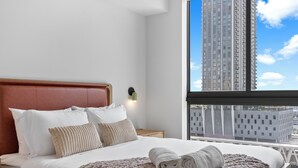 Ein Hotelzimmer mit Bett, Kissen, einer Strickdecke, einem Nachttisch, einer Wandlampe und einem Fenster mit Blick auf die Stadt.