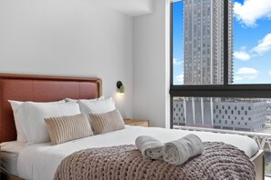 Quarto de hotel com cama, almofadas, manta de tricô, mesa de cabeceira, luminária fixada na parede e janela com vista para a cidade.