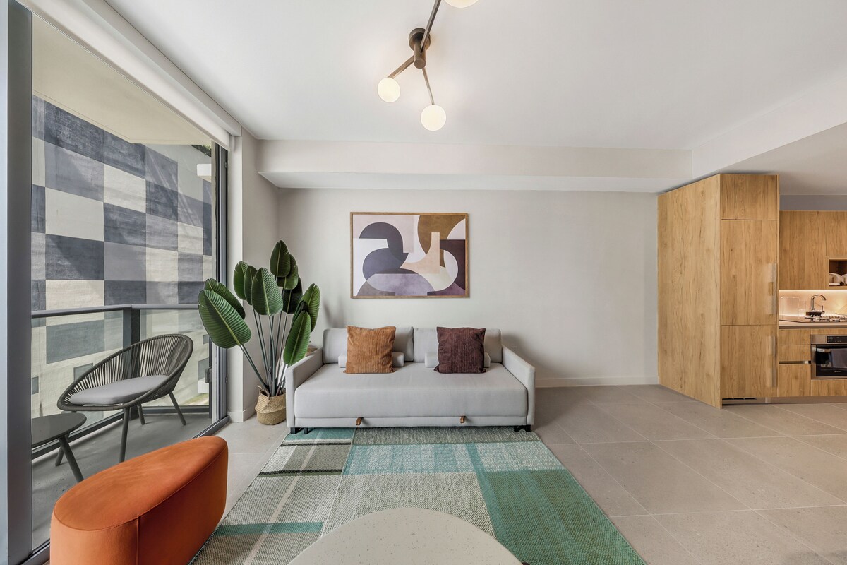 Ein modernes Wohnzimmer mit Sofa, Esstisch, Stuhl, einer großen Pflanze und einer Küchenzeile.