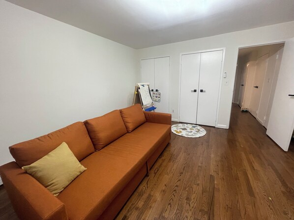 A living room with a brown sofa, white walls, wooden flooring, and a white door in the background.