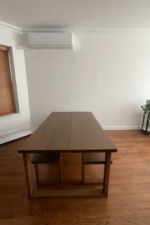 Una mesa rectangular de madera con repisa inferior, ubicada en una habitación con piso de madera y paredes blancas.