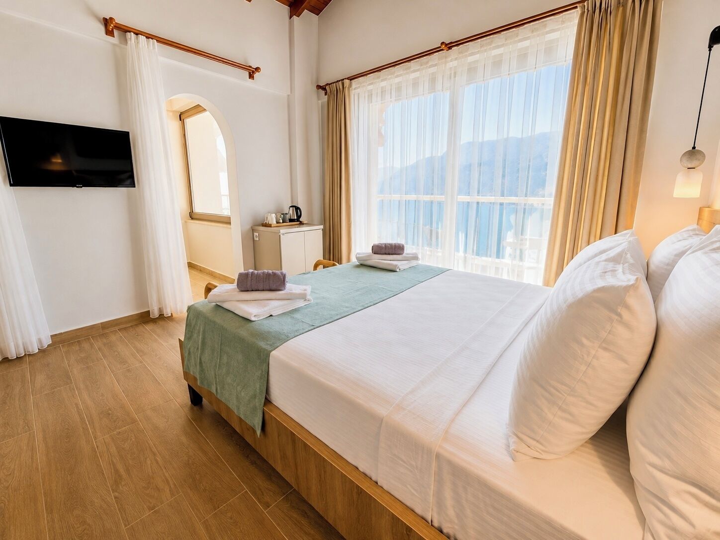 A hotel room with a bed, a TV, a desk with a kettle, and a window with curtains.