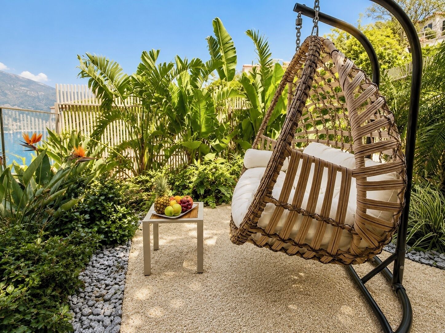 A hanging chair with a cushioned seat and back, a small table with a fruit bowl, and a garden with tropical plants and a view of a mountain.