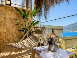 Una terraza con muro de piedra, una silla de mimbre, una mesa metálica con una botella y vasos, y vistas al mar y a las montañas.
