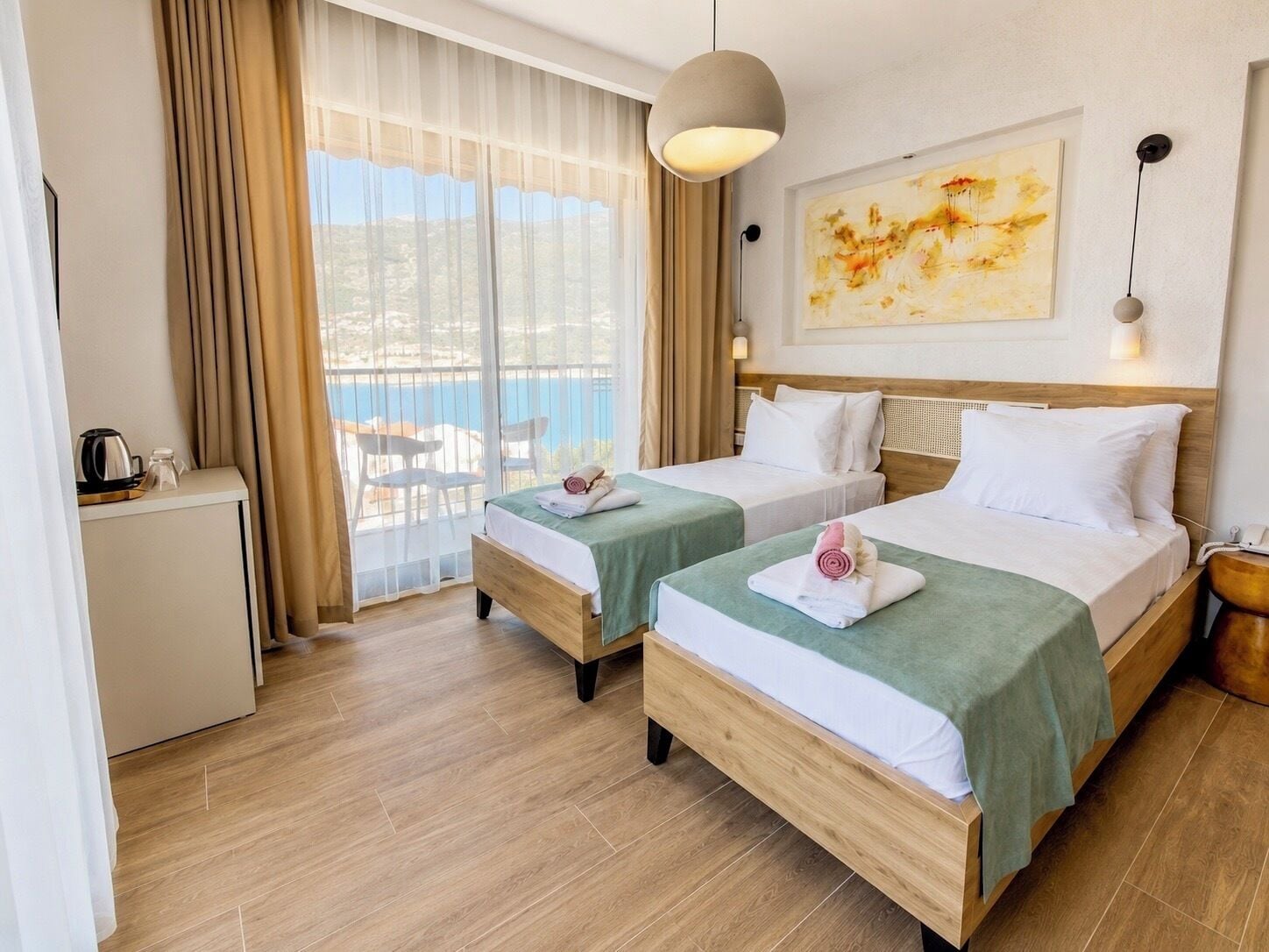 A hotel room with two beds, a view of the sea, a painting on the wall, and a small desk with a kettle.