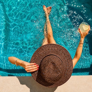Una persona relajándose en una piscina con una bebida, usando un sombrero y agarrándose del borde.