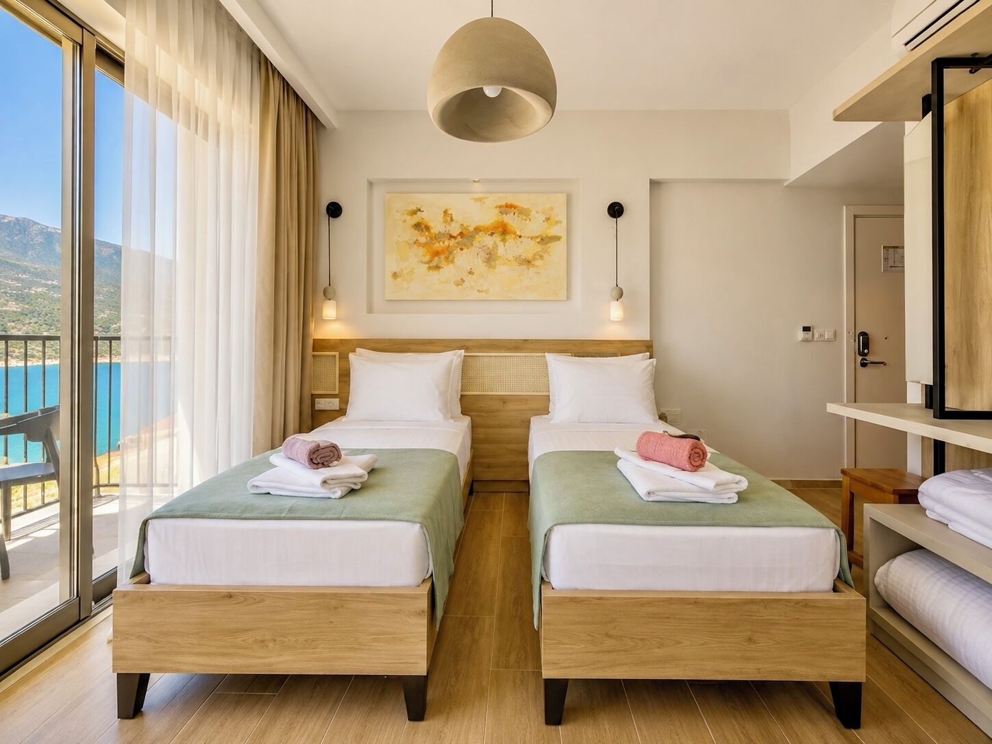 A hotel room with two beds, a large window, a painting on the wall, and a balcony with a view of the sea.