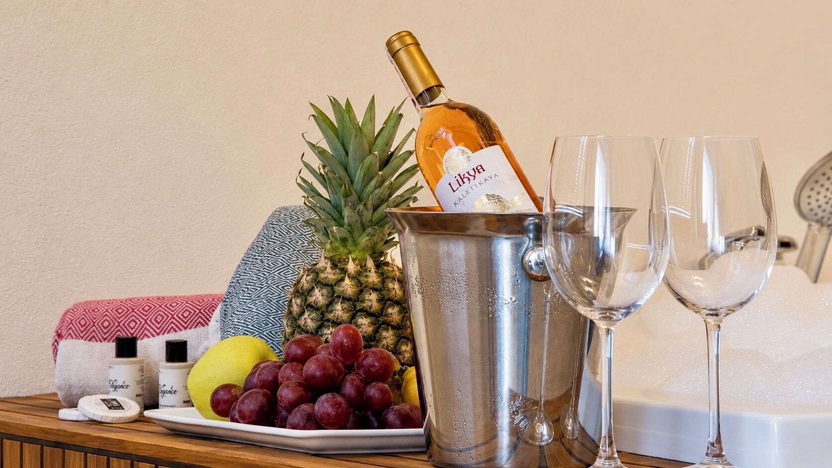 Una botella de vino Likkya Kalelikaya, una piña, uvas y dos copas sobre una mesa de madera.