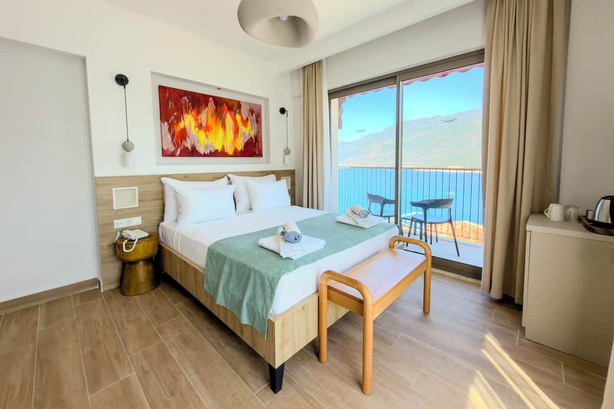 Habitación de hotel con cama, mesita de noche, silla, cuadro, ventana con vistas y balcón.
