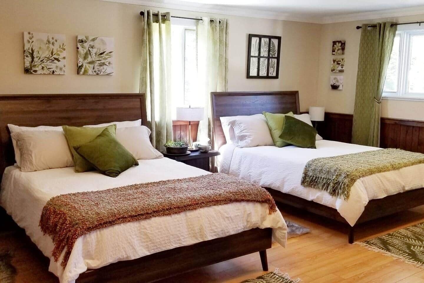 A hotel room with two beds, each with a wooden headboard, white bedding, and green pillows. There are two bedside tables with lamps, a window with curtains, and wall art.