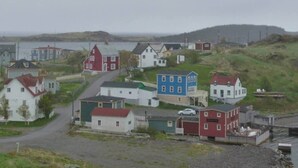 En kustby med hus längs en väg, en liten brygga och en kuperad bakgrund.