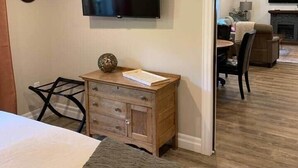 A hotel room with a bed, a wooden dresser, a television mounted on the wall, and a view into the adjacent living area. - The Trinity Guest Suite, Trinity Guest House (Trinity)