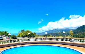 Une piscine entourée d’une clôture blanche et bordée de lampadaires, avec une végétation luxuriante et des montagnes en arrière-plan.