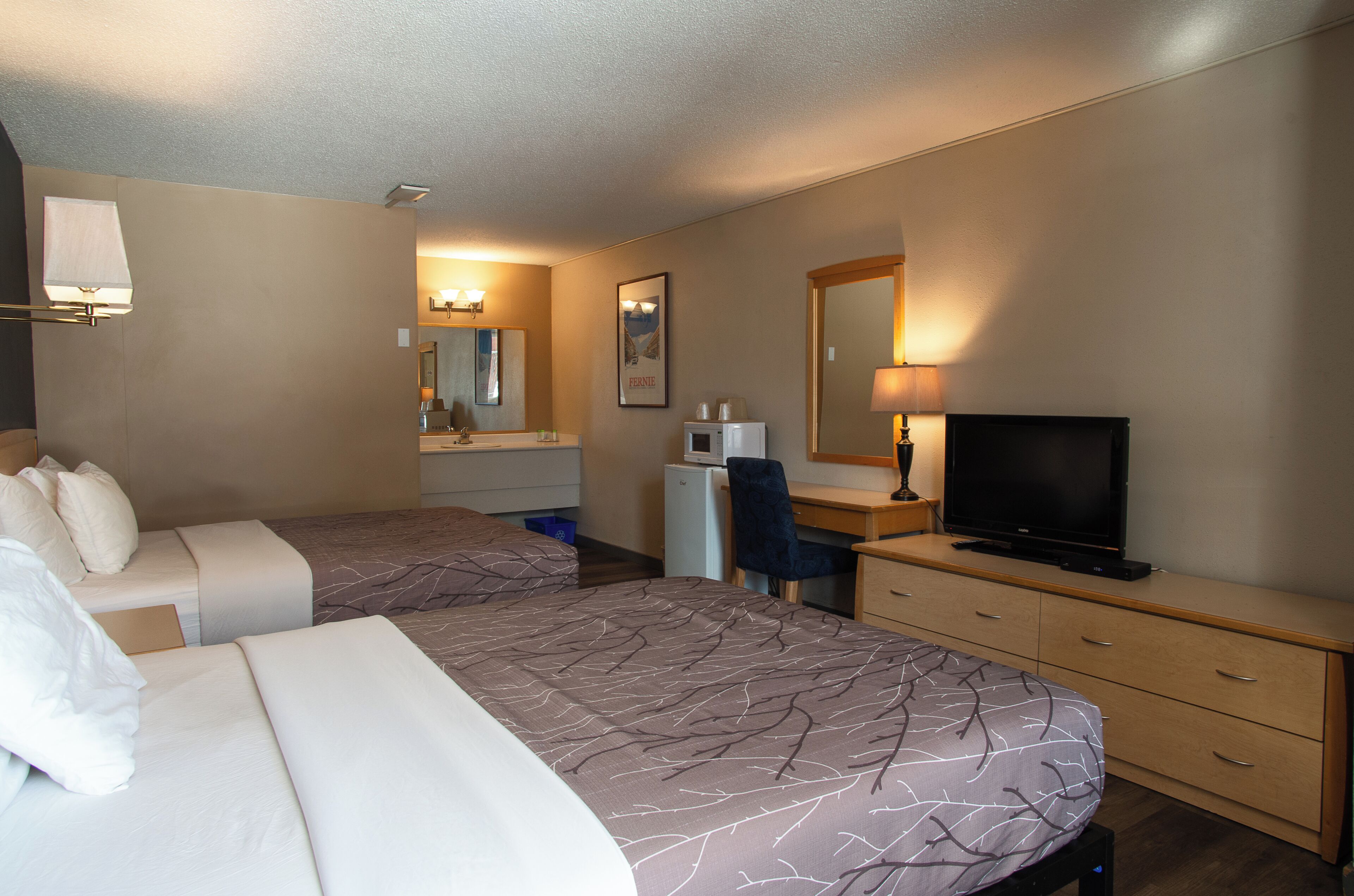 A hotel room with two beds, a desk, a television, a mirror, and a bathroom with a sink and mirror.