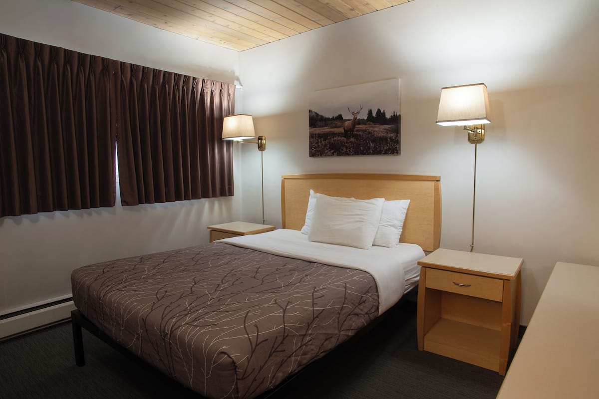 Habitación de hotel con una cama, dos mesitas de noche, una lámpara, una ventana con cortinas y un cuadro colgado en la pared.