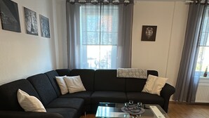 A modern living room with a black sectional sofa, a glass coffee table, and a large window with curtains. - FAMILY-FRIENDLY VACATION APARTMENT IN THE CENTER OF BRAUNLAGE (Braunlage)