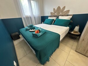A neatly made bed with teal bedding, a decorative wall headboard, a small bedside table with a lamp, and a window with sheer curtains. - Koly'Dream - Ti Kokoon - Villa with Pool in Saint-François (ST FRANCOIS)
