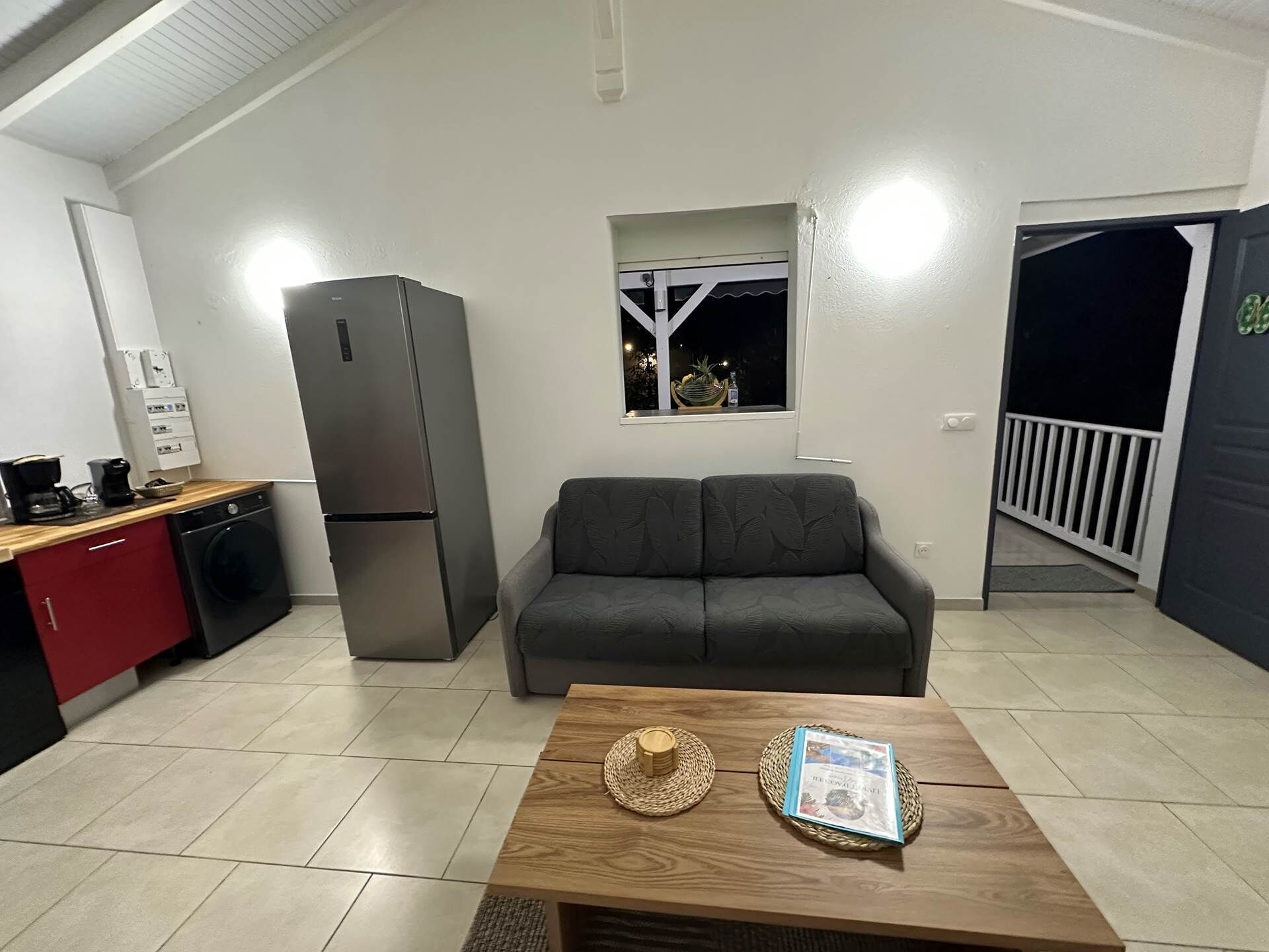 Ein kompakter Wohnraum mit einer Küchenzeile, einem grauen Sofa, einem Holztisch und einem kleinen Fenster mit Blick.