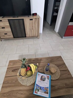 Ein Zimmer mit einer Holztruhe, einem Fernseher, einer Uhr, einem Couchtisch mit Früchten und einer Zeitschrift sowie einem gewebten Korb.