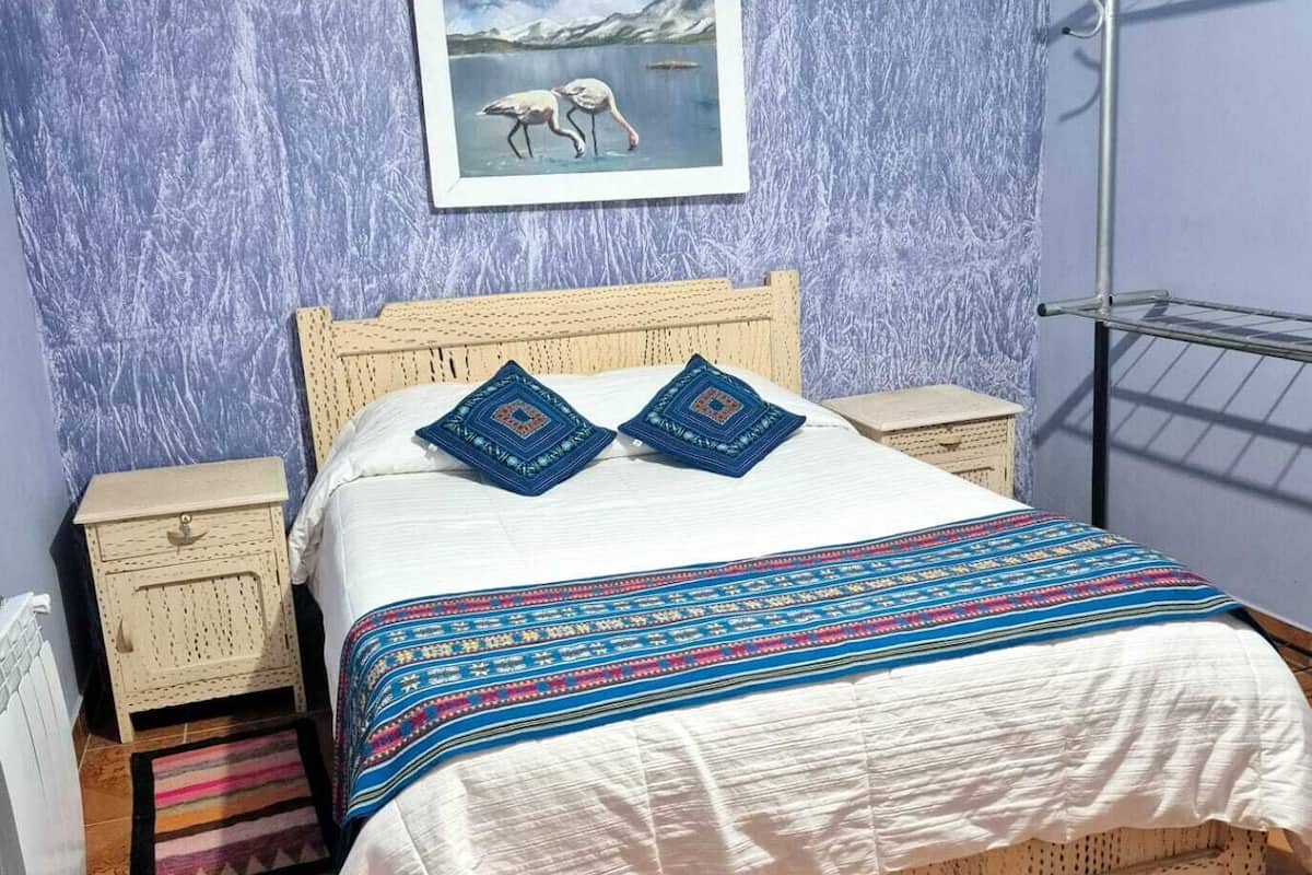 A bed with a patterned bedspread, two pillows, a wooden headboard, a nightstand, a radiator, and a framed picture on the wall.