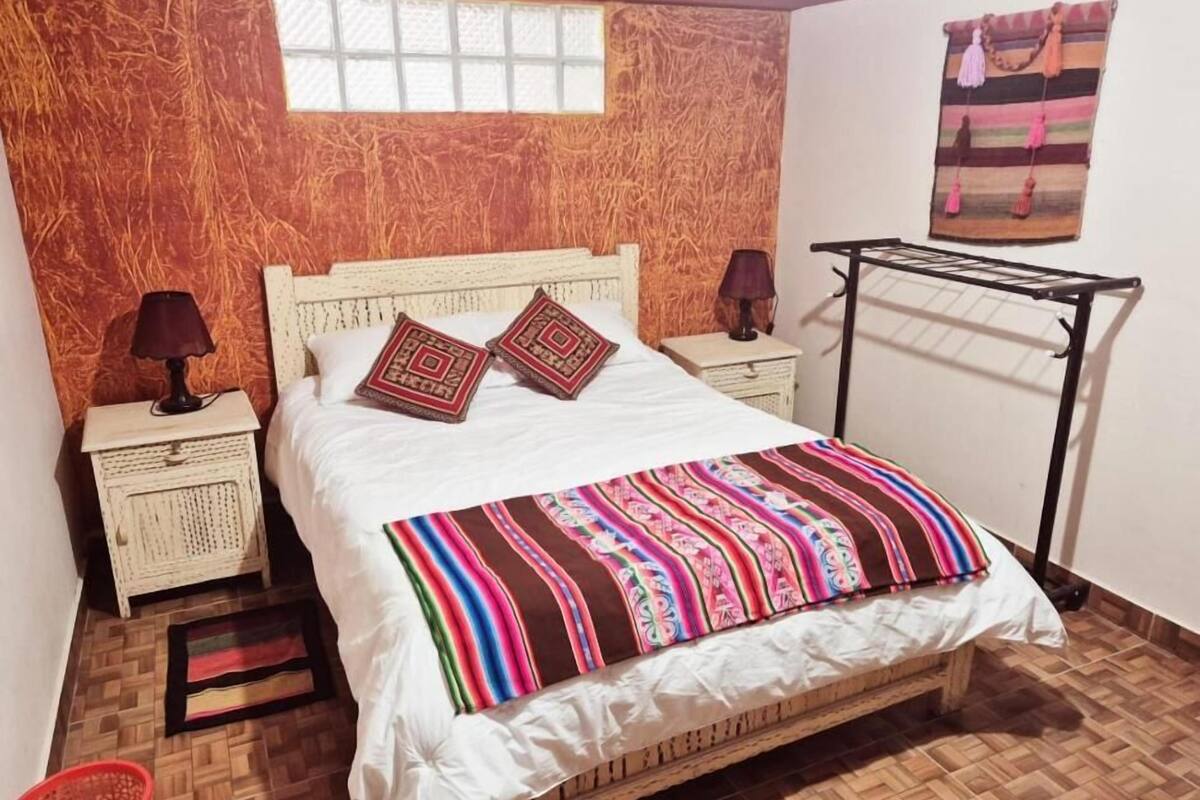 A bed with a colorful striped blanket, two decorative pillows, a nightstand with a lamp, and a wall-mounted drying rack.