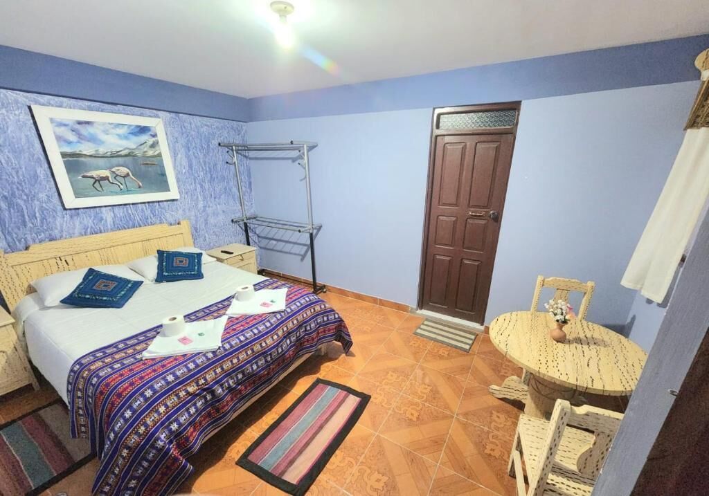 Un dormitorio con cama, una silla, una mesita con un florero, una puerta y un perchero.