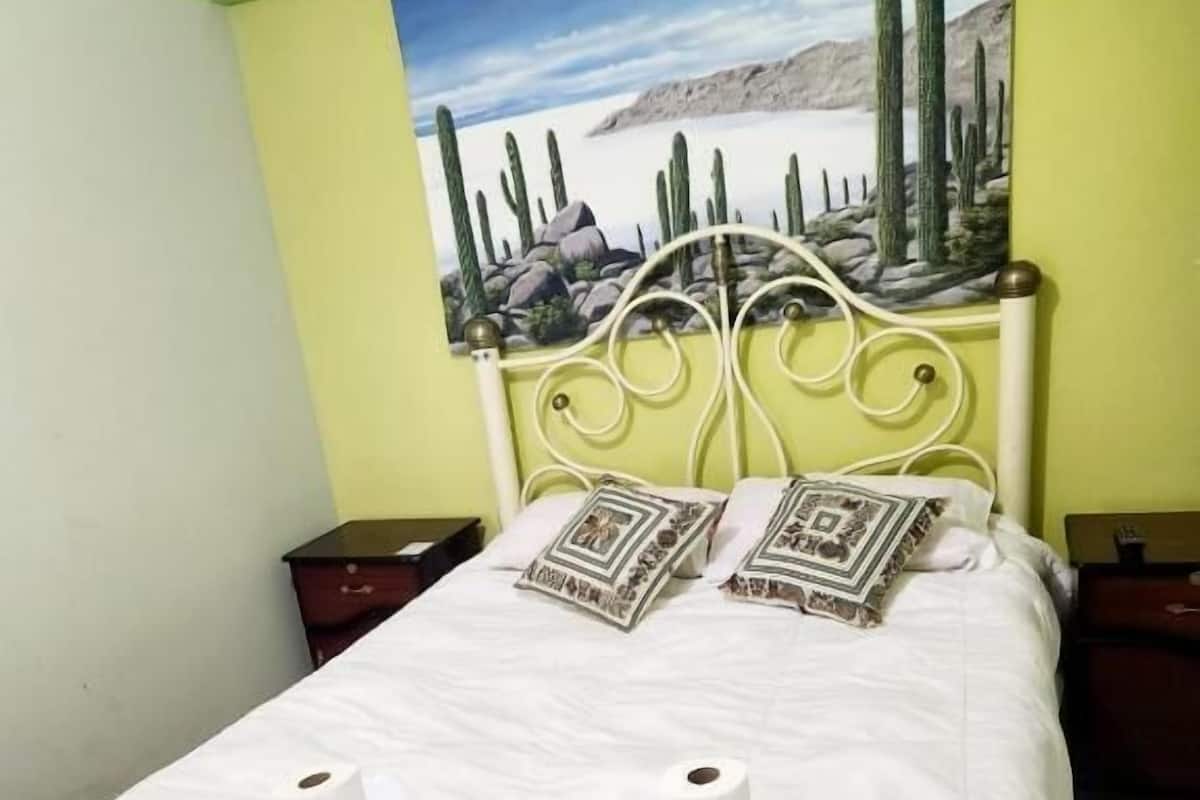 A neatly made bed with white linens and decorative pillows, a painting of cacti on the wall, and a green accent wall.