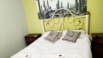 Cama bem arrumada com lençóis brancos e almofadas decorativas, um quadro de cactos na parede e uma parede de destaque verde.