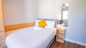 Siististi pedattu sänky, jossa on valkoiset vuodevaatteet ja yksi keltainen tyyny, yöpöytä lampun kanssa ja peili seinällä.