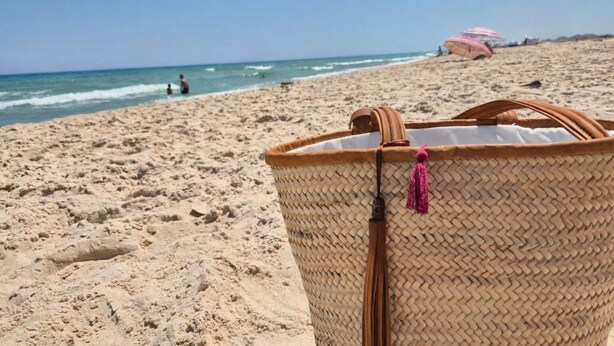 En strandscene med en flettet kurv i forgrunden og en pink parasol i baggrunden.
