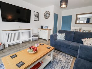 A modern living room with a blue sofa, a wooden coffee table with fruit, a white cabinet, a TV, and a clock on the wall. - Park View (Weymouth)
