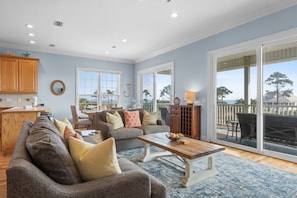A spacious living room with a large sofa, a coffee table, and a dining area. There is a sliding glass door leading to a balcony with outdoor furniture. - Blue Palms- 4/3 Gulf View home, WIFI, great views! (Cape San Blas)