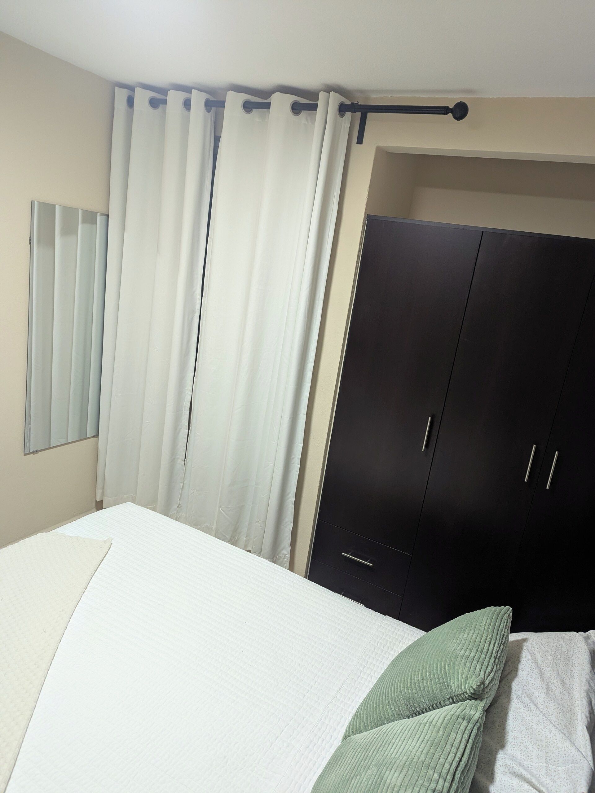 A bedroom with a bed, white curtains, a dark wardrobe, and a window with blinds.