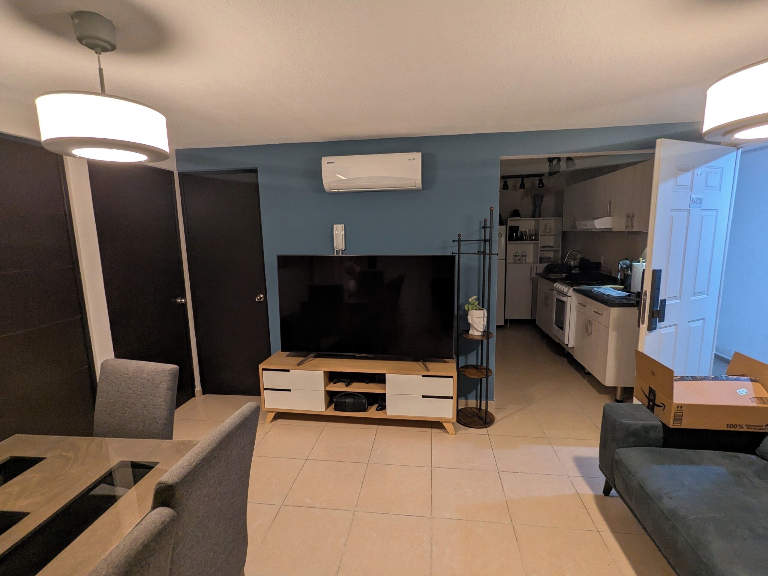 A modern living room with a flat-screen TV, a dining area with a table and chairs, a kitchen with white cabinets, and a hallway leading to another room.
