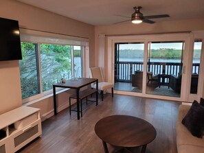 Et værelse med fjernsyn, bord, sofa og balkon med udsigt over vandet.