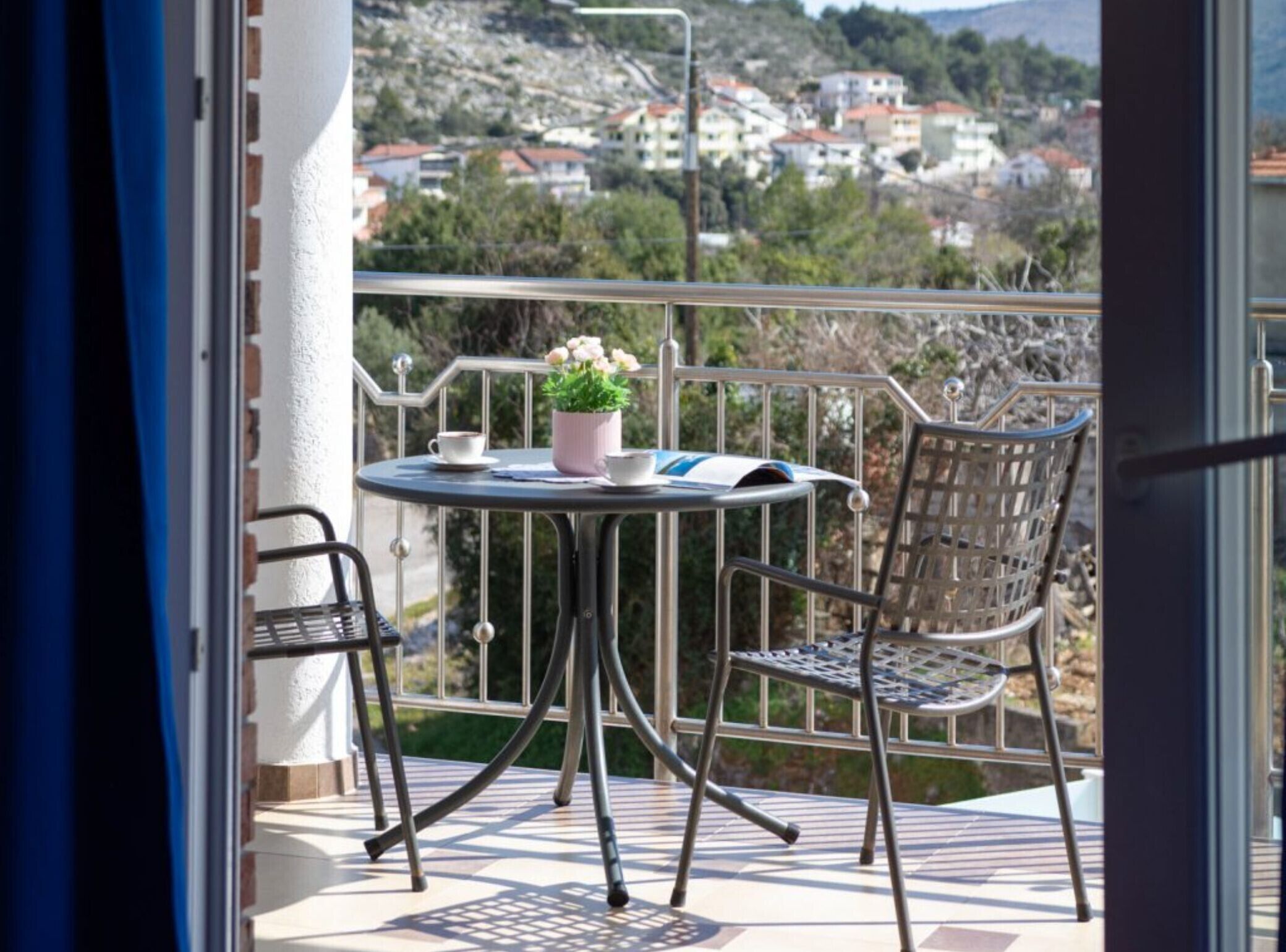 Un balcon avec une table ronde et deux chaises, un pot de fleurs et une vue sur une ville située sur une colline.
