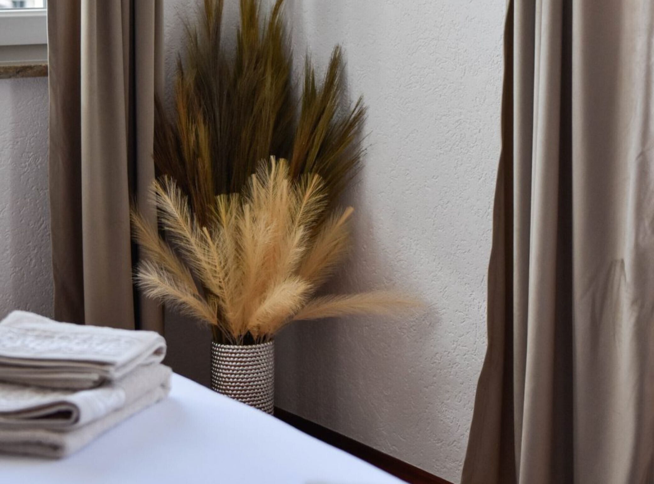 A window with beige curtains, a white wall, and a small woven basket containing dried pampas grass.