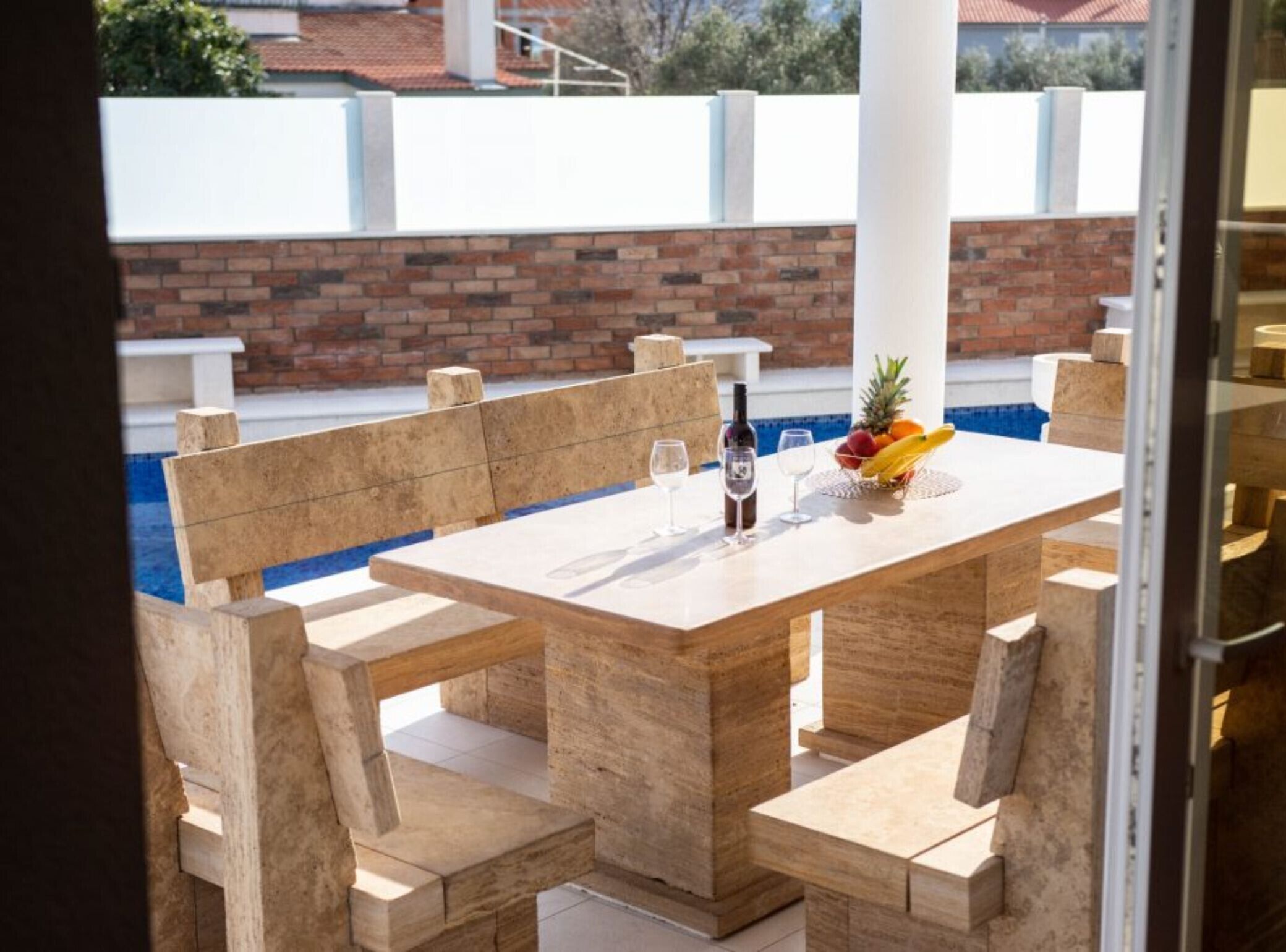 Un espace repas aménagé au bord de la piscine, avec une table et des bancs en bois, une bouteille de vin et un panier de fruits.