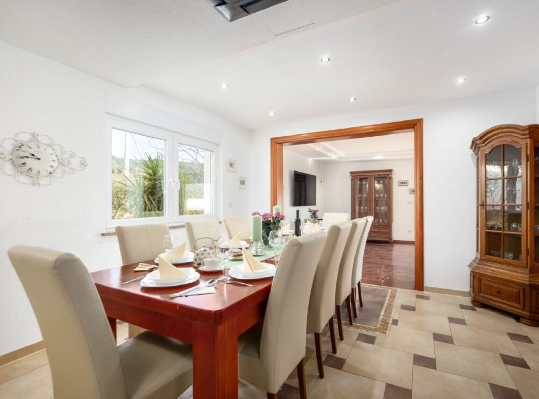 Un comedor con una mesa de madera dispuesta para cuatro personas, rodeada de sillas. A la derecha, se encuentra un mueble de madera grande con puertas de vidrio.