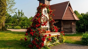 Una capilla de madera rústica con un campanario, rodeada de abundante follaje y flores rojas.