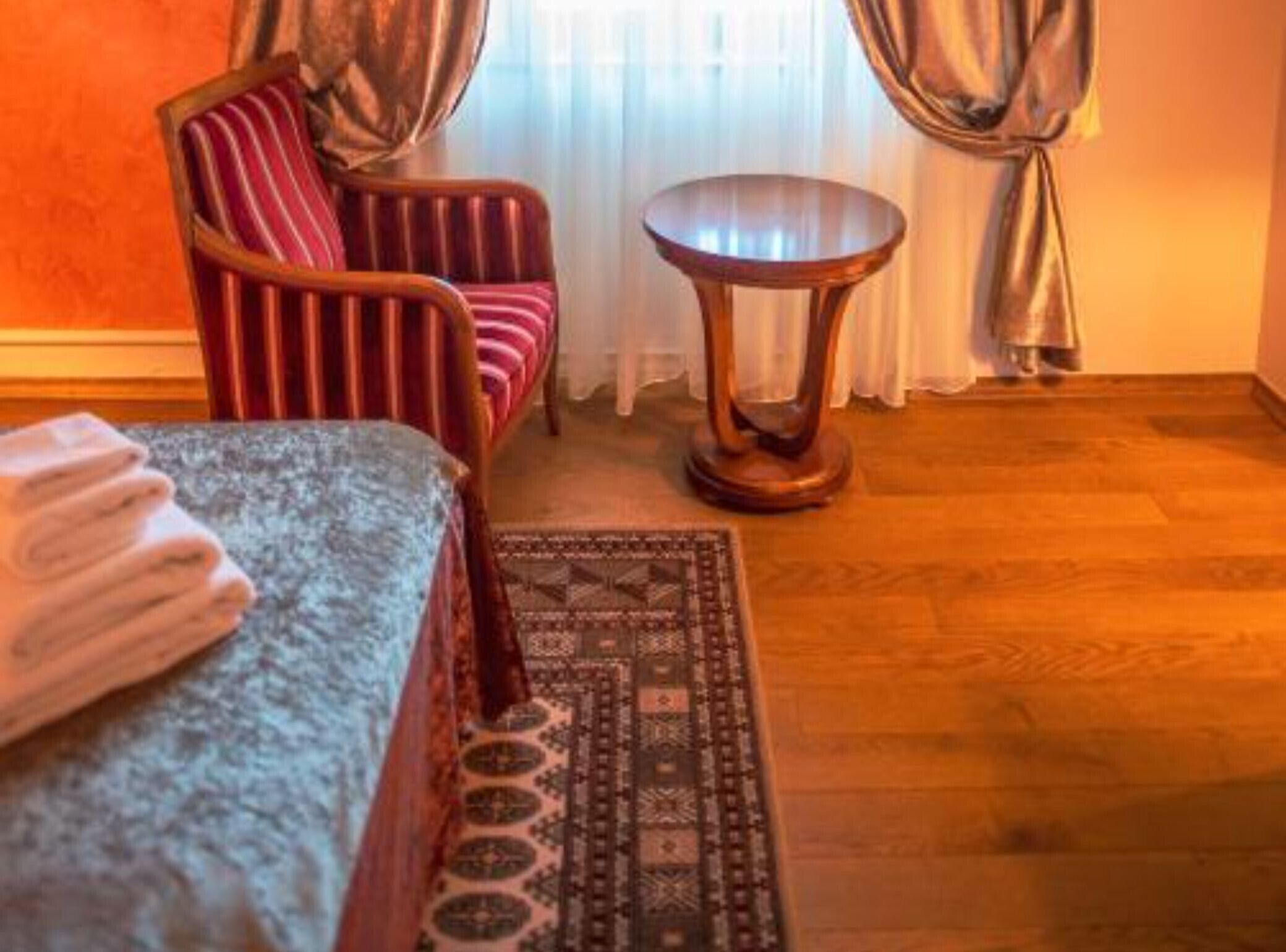 Quarto de hotel com cama, poltrona listrada, mesa redonda pequena e tapete estampado.