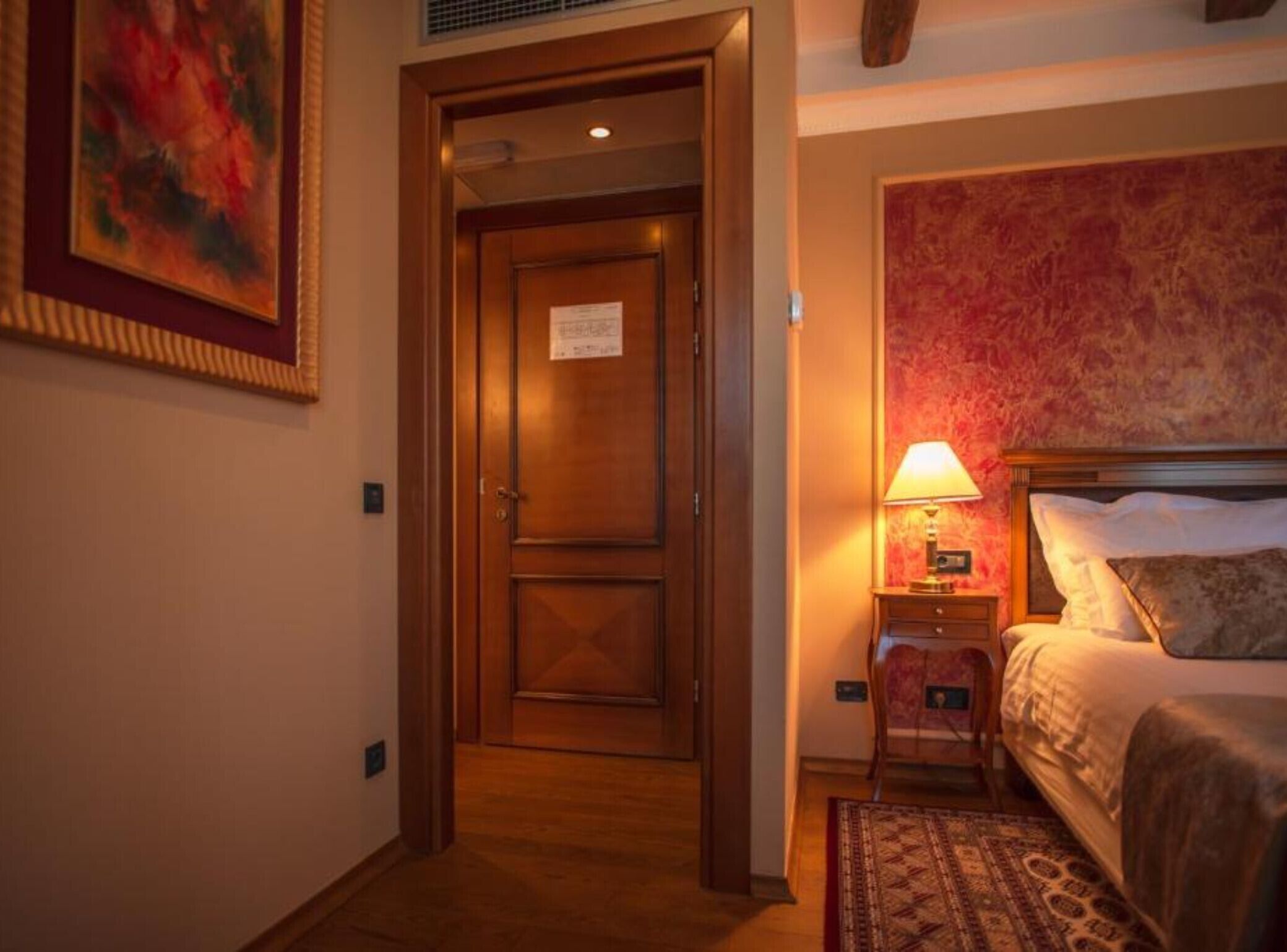 Une chambre d’hôtel avec une porte en bois, un lit doté d’une tête de lit à motifs, une table de chevet avec une lampe et un petit tapis.