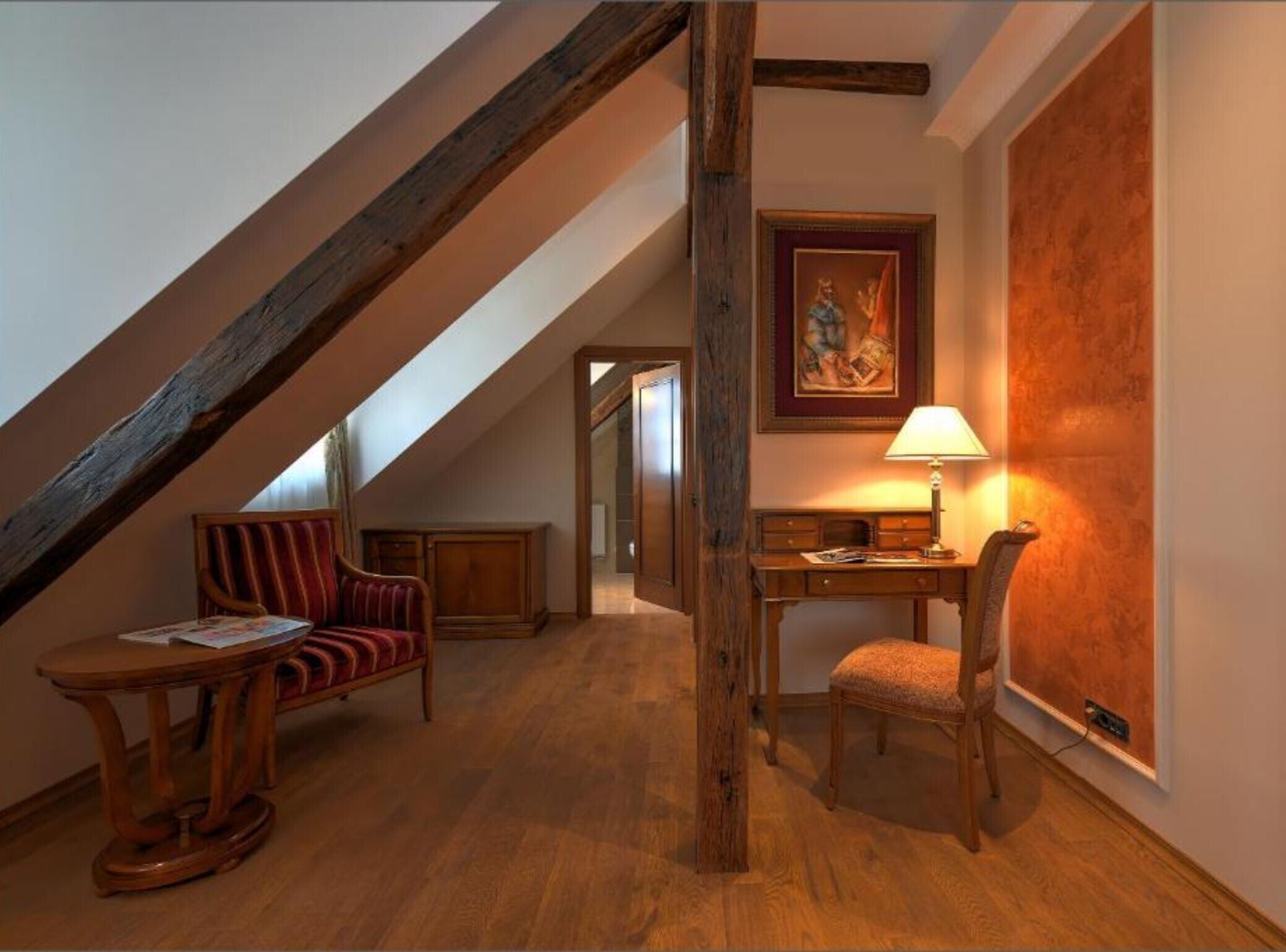 A cozy attic room with a sloped ceiling, wooden beams, a red upholstered chair, a wooden desk with a lamp, and a large framed painting on the wall.