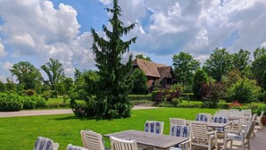 An outdoor seating area with white and blue striped chairs and tables, a thatched-roof building in the background, and a well-maintained lawn with various trees and shrubs. - Jurjevec Castle - Standard room 12 (Brezane Lekenicke)