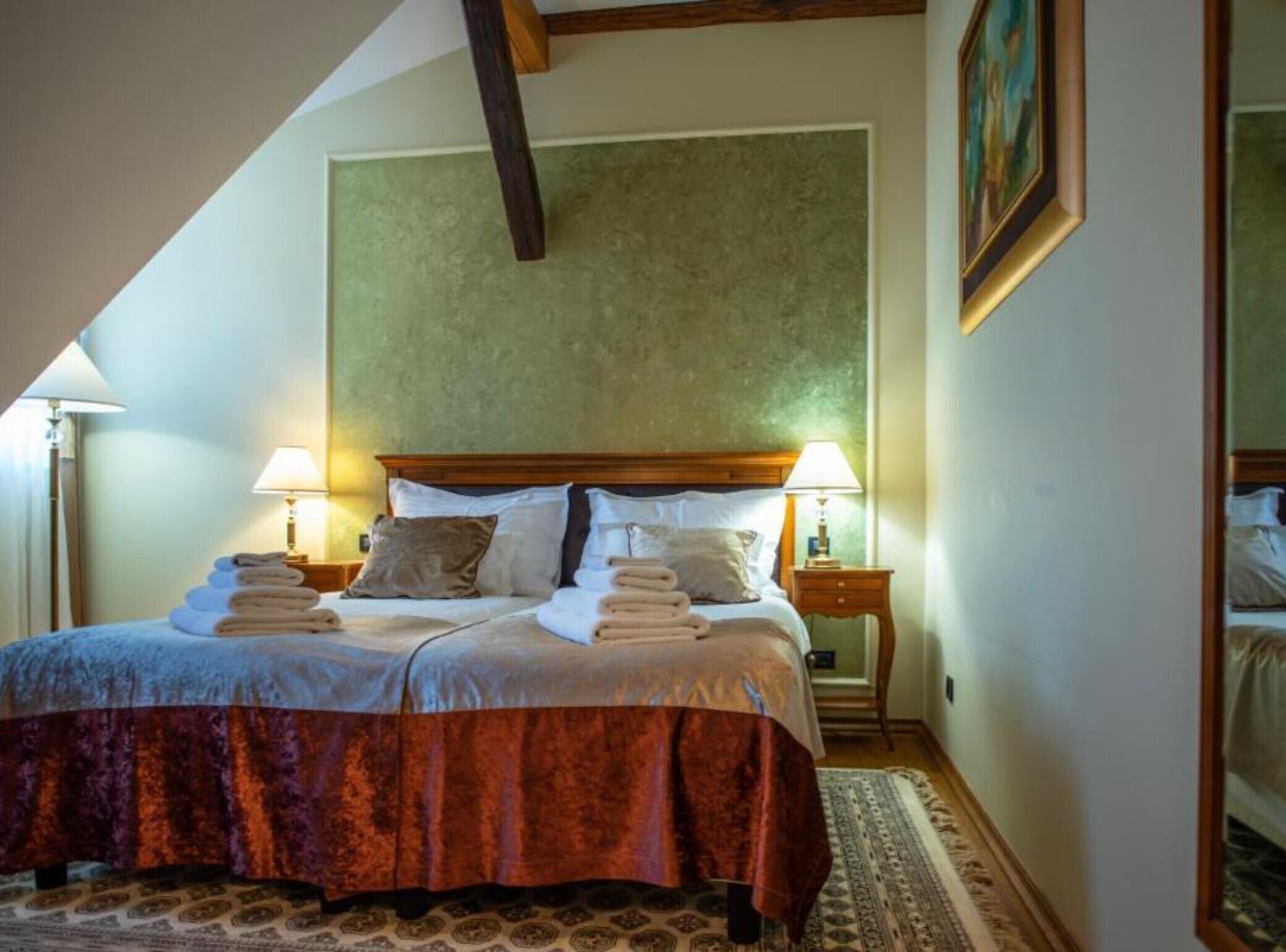 Een net opgemaakt bed met een houten hoofdeinde, twee bedlampen en een stapje gevouwen handdoeken.