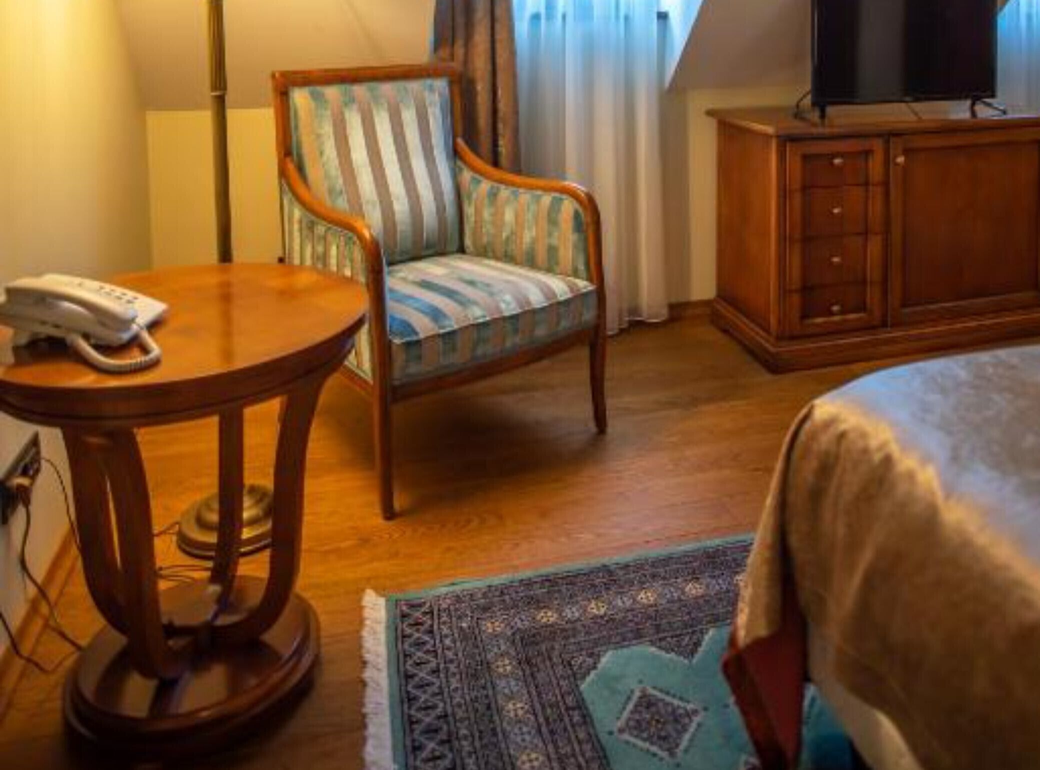 Habitación de hotel con una cama, una mesita de noche de madera con un teléfono, un sillón a rayas, un televisor en un mueble de madera y una alfombra estampada.