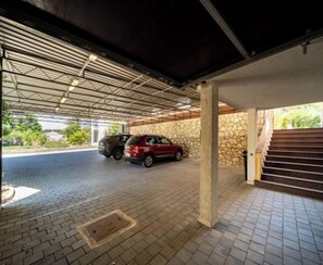 A covered parking area with two cars parked, a stone wall, and a staircase leading to an upper level. - Apartments Foric - Two bedroom apartment (1) (Rab)