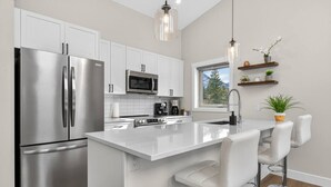 A modern kitchen with stainless steel appliances, a white island with bar stools, and a window providing a view of the outdoors. - Basecamp | Beach Access | Sports Court | Foosball (Invermere)