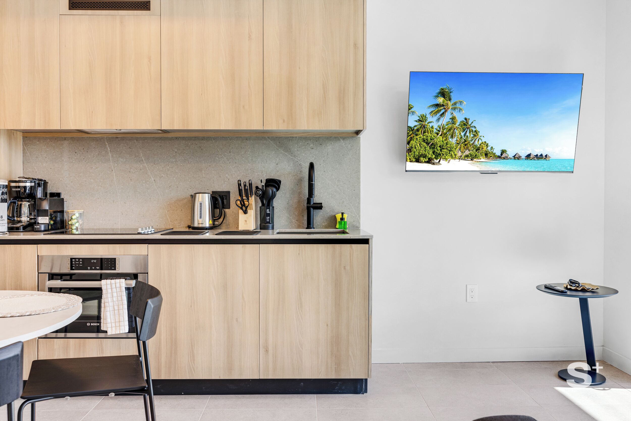 Cuina moderna amb forn integrat, cafetera i rentapesos. Hi ha una taula rodona amb una cadira i una taula rodona petita amb una cadira. Un televisor de pantalla plana muntat a la paret mostra una platja tropical.