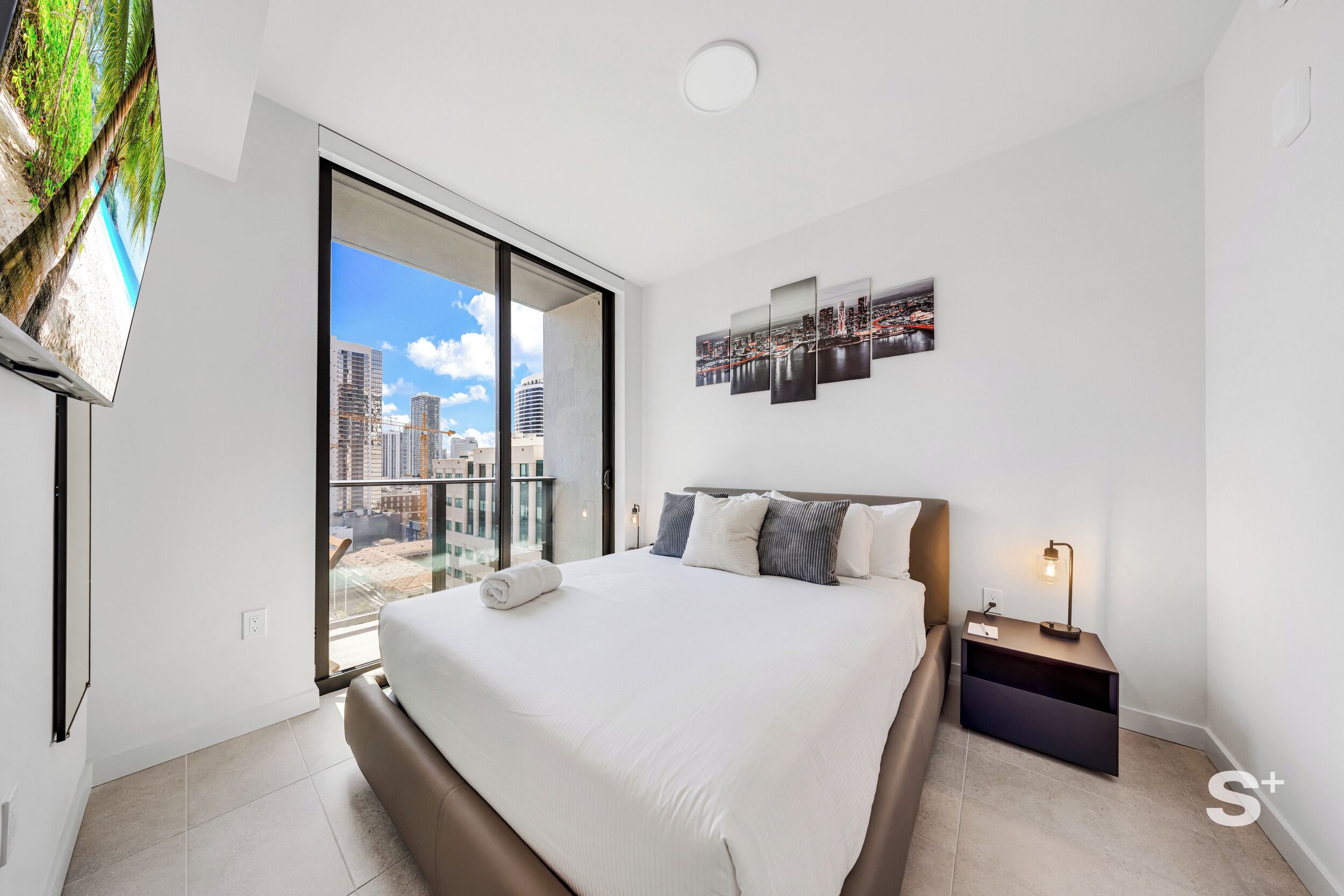Ein modernes Hotelzimmer mit einem großen Bett, einem Nachttisch, einem Flachbildfernseher und Blick auf die Stadt durch das Fenster.